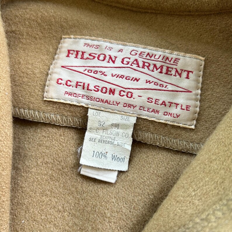 K&ouml;nnte beinhalten: Ein beigefarbenes Kleidungsst&uuml;ck aus Wolle mit einem wei&szlig;en rechteckigen Etikett mit rotem Text THIS IS A GENUINE FILSON GARMENT 100% VIRGIN WOOL C.C. FILSON CO. SEATTLE PROFESSIONALLY DRY CLEAN ONLY und einem kleineren Etikett f&uuml;r Gr&ouml;&szlig;e 32 SM und 100% Wool.
