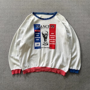 May include: A white sweatshirt with blue and red trim, featuring a graphic for the 1998 FIFA World Cup in France. The design includes the text "FRANCE 98" and a trophy image, along with the names of the host cities.