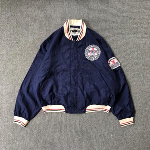 May include: A navy blue bomber jacket featuring a cream-colored ribbed collar, cuffs, and hem with red and navy stripes. The chest and sleeve display white circular patches with red and black text including TOM'S RACING and the number 36.