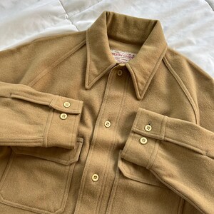 K&ouml;nnte beinhalten: Eine beigefarbene Langarm-Hemdjacke mit spitzem Kragen, durchgehender Knopfleiste und zwei Brusttaschen. Das Kleidungsst&uuml;ck hat Raglan&auml;rmel mit gekn&ouml;pften Manschetten und ein Etikett am inneren Halsausschnitt mit der Aufschrift Filson Garment.