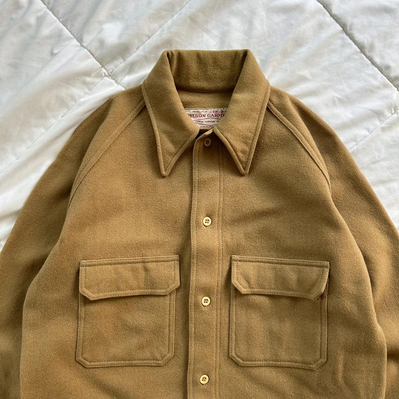 K&ouml;nnte beinhalten: Ein beigefarbenes Langarm-Hemd mit spitzem Kragen, Knopfleiste auf der Vorderseite und zwei Brusttaschen mit Klappen. Das Etikett am Innenkragen zeigt den Schriftzug "FILSON GARMENT" in roten Buchstaben auf wei&szlig;em Hintergrund.