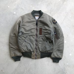 May include: An olive green bomber jacket featuring a front zipper closure, ribbed collar, cuffs, and hem. The jacket includes two front snap-button pockets, a rectangular brown patch on the chest, and a circular emblem on the upper left sleeve.