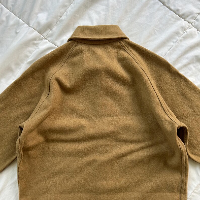 K&ouml;nnte beinhalten: Eine hellbraune Jacke mit klassischem Umlegekragen, langen &Auml;rmeln mit Raglann&auml;hten und einer glatten, filzartigen Textur. Das Kleidungsst&uuml;ck ist von hinten abgebildet und zeigt die klaren Linien sowie den einheitlichen, kamelbraunen Farbton.