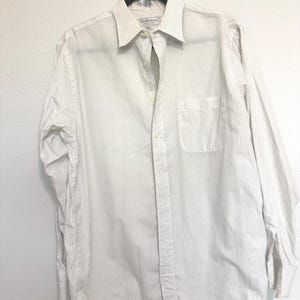 以下が含まれることがあります： 細い灰色の縦縞模様の白いシャツ。胸ポケットと長袖が付いています。襟にはブランド名「Burberrys」が見えます。