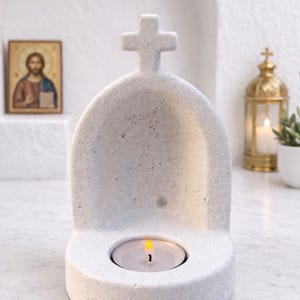 May include: A white stone candle holder, topped with a cross. The holder features an arched design and a space for a tea light candle. A lit candle is inside, emitting a warm glow. The background includes a religious icon and a decorative lantern.