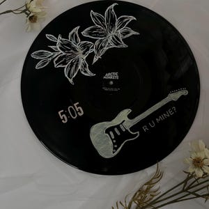 May include: A black vinyl record featuring white hand-painted illustrations of two lilies, an electric guitar, the text 5:05, and the text R U MINE?. The centre label displays the text ARCTIC MONKEYS and the song title WHY'D YOU ONLY CALL ME WHEN YOU'RE HIGH.