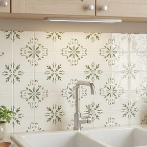 May include: A kitchen scene featuring a white double sink with a chrome faucet. The backsplash is tiled with a white background and a green floral pattern. Above the sink are light-colored cabinets and a small light fixture.