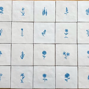 May include: A collection of twenty-five white square tiles, each featuring a different blue floral illustration. The designs include roses, tulips, and other stylized flowers. The tiles are arranged in a grid pattern on a light brown surface.