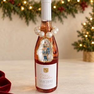 Könnte beinhalten: Eine Flasche Comte de la Chevalière Sancerre Wein mit einer weißen Perlenkette um den Flaschenhals und einem Austernschalen-Ornament, das mit einer kleinen blau-weißen Nussknacker-Figur auf der Vorderseite verziert ist.