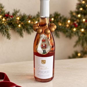 Könnte beinhalten: Eine Glasflasche mit Comte de la Chevalière Sancerre Wein und einem weißen Etikett mit schwarzer Schrift. Der Flaschenhals ist mit einem Ring aus matten Goldperlen und einer Austernschale verziert, die eine kleine Nussknackerfigur enthält.