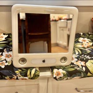 May include: Off-white iMac G3 mirror with a rectangular frame. The mirror has a built-in speaker and a small Apple logo. The mirror is resting on a floral patterned cushion.