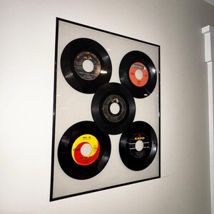 May include: Framed wall art featuring five vintage vinyl records. The records are black with colorful labels in red, orange, and yellow. The records are arranged in a rectangular frame with a black border, mounted on a white wall.