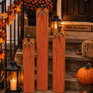 May include: Three tall, rectangular wooden blocks painted in a rustic orange finish, each topped with a dark stem, a small cluster of artificial autumn leaves, and a decorative raffia bow, arranged vertically as seasonal porch decor.