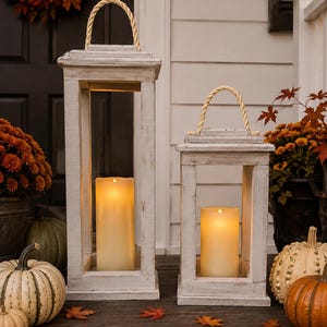 May include: Two rectangular wooden lanterns with a distressed white finish and natural rope handles. Each lantern features an open-frame design and contains a single cylindrical LED pillar candle emitting a warm, soft yellow glow.