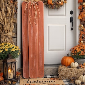 May include: A tall, vertical, orange-painted wooden pumpkin sign with a raffia bow topper, placed on a rectangular doormat featuring the word Welcome, pumpkin illustrations, and a black and white buffalo check border pattern.