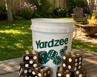 May include: A white plastic bucket with the green text Yardzee and green illustrations of dice, accompanied by five large, dark brown wooden dice featuring white circular pips on each face, arranged in a cluster on a wooden surface.