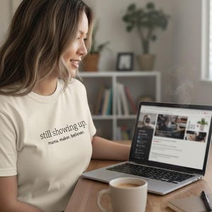 Puede incluir: Una camiseta color crema con el texto "still showing up. mama. maker. believer." es usada por una persona sentada en un escritorio de madera. Una computadora portátil, una taza blanca con café, un cuaderno y un bolígrafo están en el escritorio.