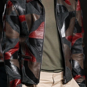 Può includere: Giacca patchwork multicolore in pelle con chiusura a zip. La giacca, nei toni del nero, grigio, marrone e rosso, è indossata sopra una maglia verde a coste e pantaloni beige. Il design presenta una varietà di trame e motivi.