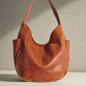 May include: A brown leather hobo bag with a curved top and two exterior pockets. The bag has a single shoulder strap and is made of a soft, textured leather. The bag is a warm, earthy tone.
