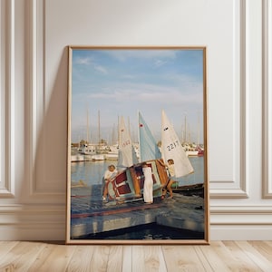 May include: A framed vertical wall art print depicting three individuals on a wooden dock manoeuvring a small sailboat with white and blue sails. The scene features a harbour with multiple moored boats under a clear blue sky, captured in a vintage style.