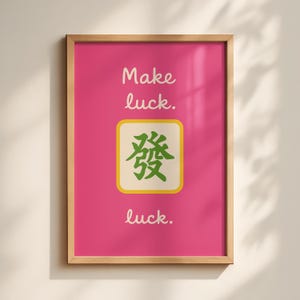Könnte beinhalten: Ein gerahmter Druck mit einem leuchtend rosa Hintergrund. Der Text "Make luck. luck." ist in weißer Schrift geschrieben. Eine quadratische Kachel mit einem grünen chinesischen Zeichen ist zentriert.