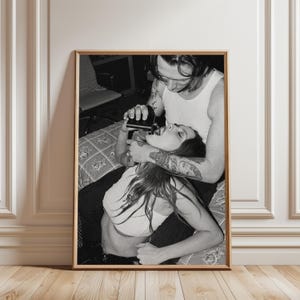 以下が含まれることがあります： 白黒の額入りウォールアートプリント。部屋にいる2人の人物が描かれています。白いタンクトップを着た人物が、白いクロップトップを着たもう一人の人物の口に、暗い色のフラスコから液体を注いでいる様子が写し出されています。