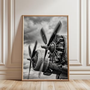 May include: A black and white framed wall art print featuring a close-up view of a vintage airplane engine with two propellers in motion. The detailed radial engine components are visible against a cloudy sky background, captured in high contrast.