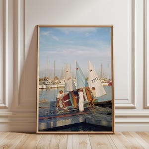 May include: A framed wall art print depicting a harbour scene with three sailboats. Two individuals in light clothing are lifting a small wooden boat onto a wooden dock. The sailboats feature white and light blue sails with the numbers 1562 and 2217.