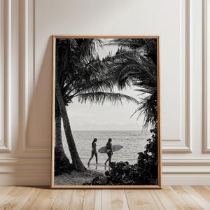 May include: A black and white framed wall art print depicting a beach scene framed by palm trees. Two figures walk along the shoreline, one carrying a surfboard toward the ocean. The composition features high contrast shadows and bright sky.