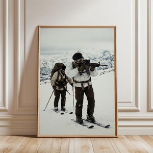 May include: A framed wall art print depicting two individuals in white and dark tactical winter gear, wearing black berets and carrying rifles while skiing across a snowy mountain landscape under a cloudy sky. The scene captures a military-style winter exercise.
