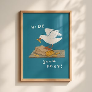 Op de afbeelding: Een ingelijste kunstprint met een witte meeuw met oranje poten die op een bruine papieren zak met goudkleurige frietjes op een houten plank staat, tegen een blauwgroene achtergrond met de handgeschreven witte tekst HIDE YOUR FRIES!