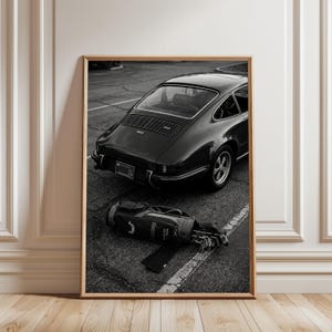 以下が含まれることがあります： アスファルトの上に駐車されたヴィンテージスポーツカーの後部を写したモノクロの額入りプリント。車の前にはゴルフバッグと数本のゴルフクラブが置かれている。車のナンバープレートとリアエンジングリルがはっきりと確認できる。