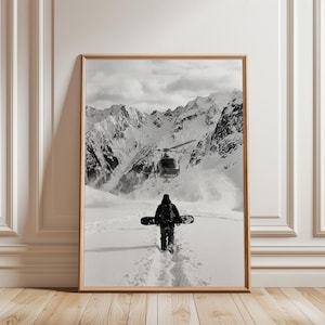 May include: A black and white framed wall art print depicting a person carrying a snowboard across a snowy landscape towards a helicopter hovering in front of jagged, snow-covered mountain peaks. The scene captures a vast, wintry outdoor environment.