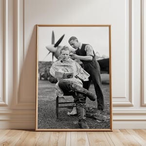 Puede incluir: Una fotografía enmarcada en blanco y negro que muestra a una persona sentada en una silla leyendo un libro mientras otra le corta el pelo. La escena se desarrolla al aire libre con una gran hélice de avión visible al fondo tras las figuras.