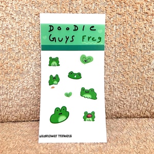 May include: A sticker sheet featuring various green frog illustrations. The top of the sheet has the text "Doodle Guys Frog" in black and green. The bottom of the sheet has the text "Wildflower Trinkets".