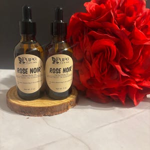 May include: Two brown glass bottles of "Rose Noir" herbal body oil with black droppers, displayed on a wooden slice. A large, vibrant red rose is in the background. Each bottle contains 57g of product.