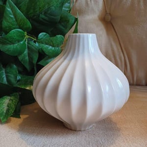 May include: A white ceramic vase with a rounded, bulbous body and a narrow neck. The surface of the vase features vertical, fluted ridges that extend from the base to the opening, creating a consistent, repeating pattern around the entire object.