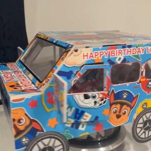 Puede incluir: Un coche de juguete azul y blanco decorado con personajes de dibujos animados y el texto "HAPPY BIRTHDAY LOGAN". El coche presenta imágenes de perros de dibujos animados, estrellas y huellas de patas. Las ruedas son negras y plateadas.