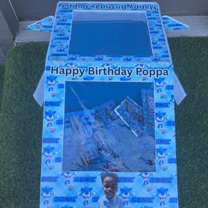 May include: A blue and white birthday gift box with the text "Happy Birthday Poppa". The box is decorated with Sonic the Hedgehog graphics and a photo of a child. Inside are wrapped candies.