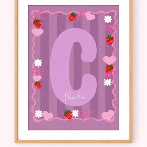 Op de afbeelding: Een ingelijste print met een grote lavendelkleurige letter "C" en de naam "Charlie". De achtergrond heeft verticale strepen. De rand bevat aardbeien, harten, strikken en bloemen in roze en wit.