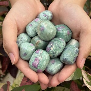 Peut inclure: Une collection de pierres polies présentant une texture lisse et brillante. Chaque pierre affiche une couleur de base vert clair marbré avec des inclusions irrégulières distinctes de dépôts minéraux magenta foncé et rouge.