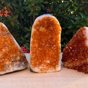May include: Three standing citrine crystal geodes with deep orange and golden yellow faceted points. The crystals feature a rough, natural outer edge and a polished base, displaying a vibrant, translucent, and sparkling crystalline structure.