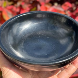 Könnte beinhalten: Eine schwarze, runde Schüssel mit glatter, polierter Oberfläche. Die Schüssel wird in einer Hand gehalten, mit einem verschwommenen Hintergrund aus rotem und grünem Laub. Das Design der Schüssel ist schlicht und elegant.