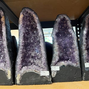 May include: Four tall amethyst geodes standing upright in a row. Each geode features a deep purple crystalline interior with jagged, pointed edges and a dark, solid base. Small white rectangular labels are attached to the front of each stone.