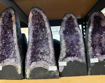 Peut inclure: Quatre grandes géodes d'améthyste disposées en rangée. Chaque géode présente un intérieur cristallin violet profond avec des bords dentelés et une base sombre et solide. De petites étiquettes rectangulaires blanches sont fixées sur chaque pierre.