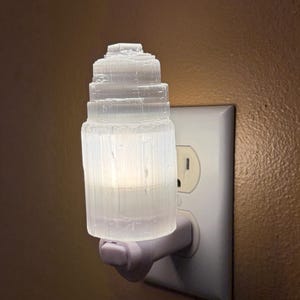 May include: A white selenite night light plugged into a wall outlet. The night light has a tiered, cylindrical design, and emits a soft, warm glow. The outlet is white, and the wall is a warm brown color.