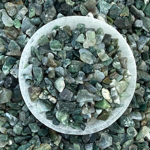 May include: A white bowl filled with small, irregularly shaped green and gray stones. The stones are a mix of light and dark shades, creating a natural, textured appearance. The bowl sits on a bed of similar stones.