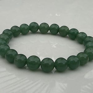 May include: A circular stretch bracelet composed of numerous smooth, round, dark green beads. The beads are uniform in shape and size, featuring a polished, glossy finish that reflects light across their spherical surfaces, arranged in a continuous loop.