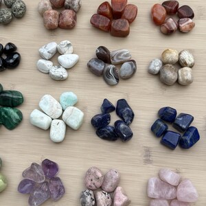 Könnte beinhalten: Sortiment polierter Edelsteine in verschiedenen Farben, darunter grüner Malachit, blauer Lapislazuli und rosa Rosenquarz. Die Steine sind auf einer hellen Holzoberfläche angeordnet und zeigen ihre natürlichen Texturen und Farbtöne.