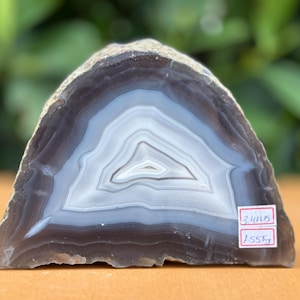 May include: A polished agate slice featuring concentric bands of dark gray, light gray, and white. The stone has a triangular shape with a rough outer edge. A small white label on the lower right displays the text 3.4 lbs and 1.55 kg.